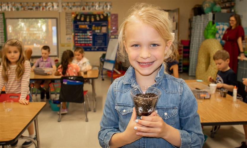 A blonde girl smiles while holding a small plastic cup full of dirt.
